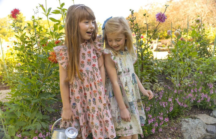 Two young girls in floral dresses standing in a garden with flowers and plants.