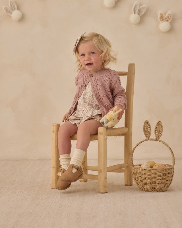 Young girl sitting on a wooden chair holding a teddy bear, with a basket and decorative elements in the background.