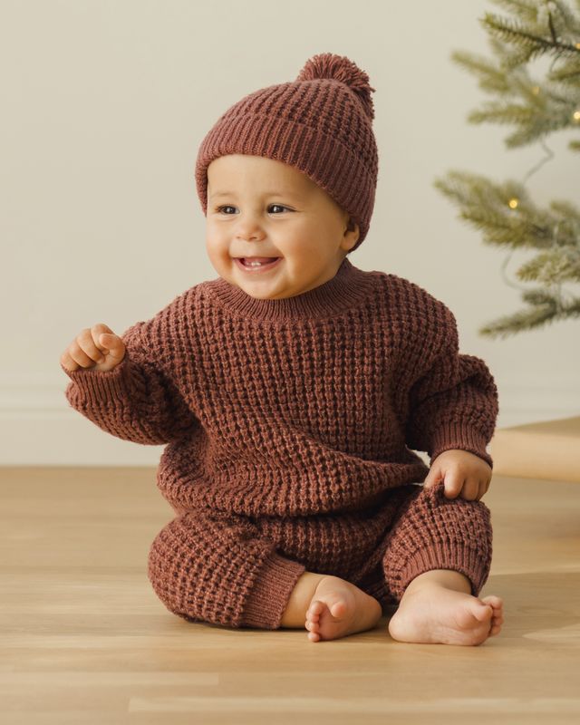 Baby wearing a cranberry knitted outfit and hat, sitting on a wooden floor with a Christmas tree in the background.