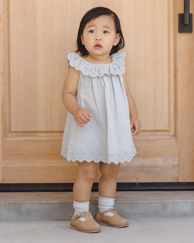 Young child wearing a light blue dress with ruffled details, standing in front of a wooden door.