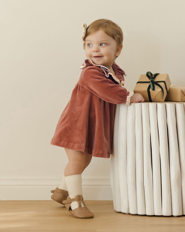 Child in a rust-colored dress standing next to a white radiator with wrapped gifts on a wooden floor.