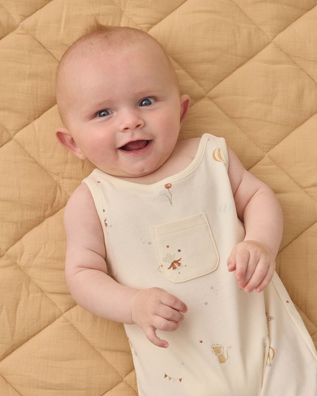 Baby wearing a light-colored outfit with cartoon carnival designs with a pocket, lying on a quilted beige surface.