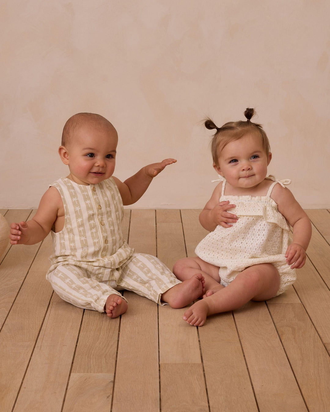 two babies sitting in rylee and cru outfits. one in palm stripe jumpsuit and the other in an ivory eyelet bubble. 