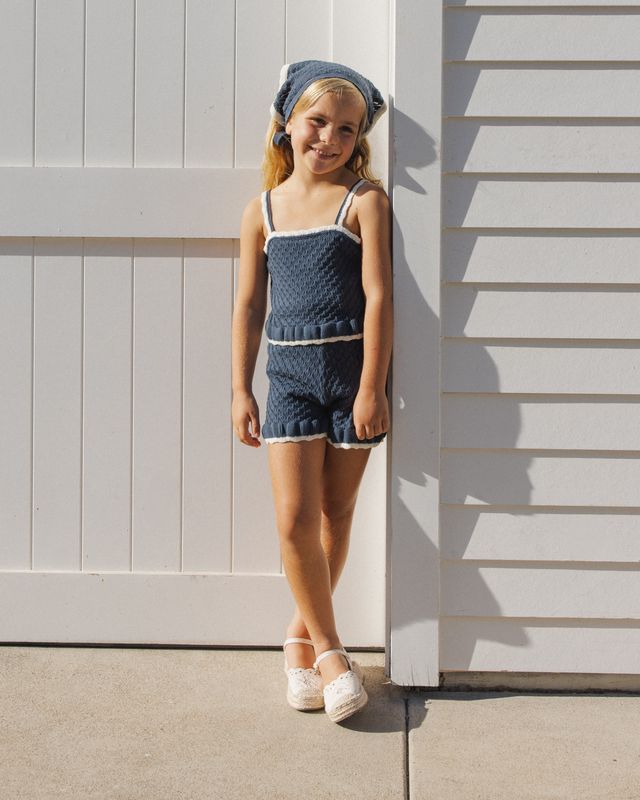 Young girl in a blue knit outfit standing against a white wall.