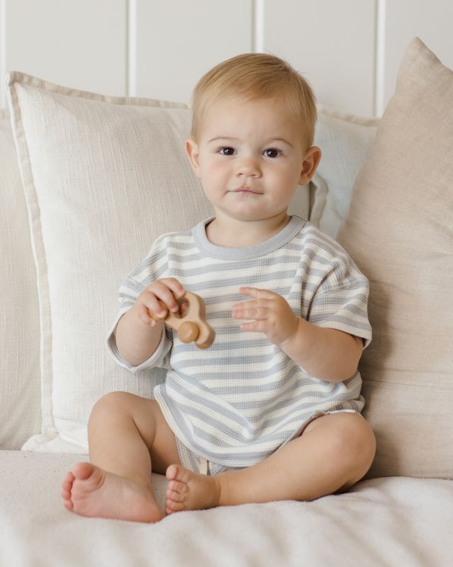 Baby sitting on a couch holding a wooden toy, wearing a striped onesie.