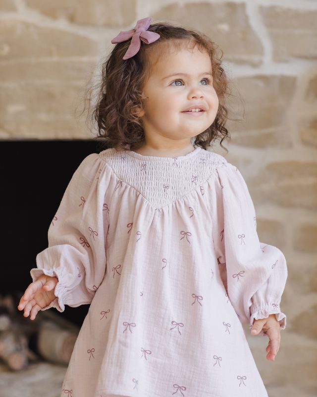 Child wearing a light pink dress with bow patterns in front of a stone wall.