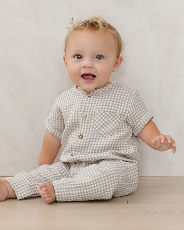 Baby wearing a sage checkered outfit sitting on a light wooden floor.