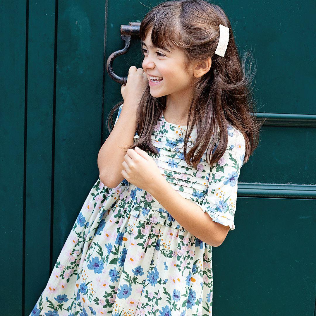 young girl modeling the ivory dress with blue and lavender flowers with pops of orange. the girl is laughing and the wind is blowing. 