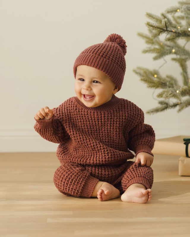 Baby wearing a cranberry knitted outfit and hat sitting on a wooden floor with a Christmas tree in the background.