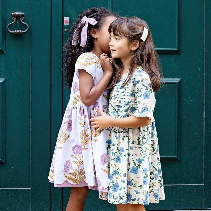 two young girls wearing pink chicken dresses telling secrets in front of a beautiful green door. one girl in the blue floral dress and one in the white dress with lavender bohemian flowers. 