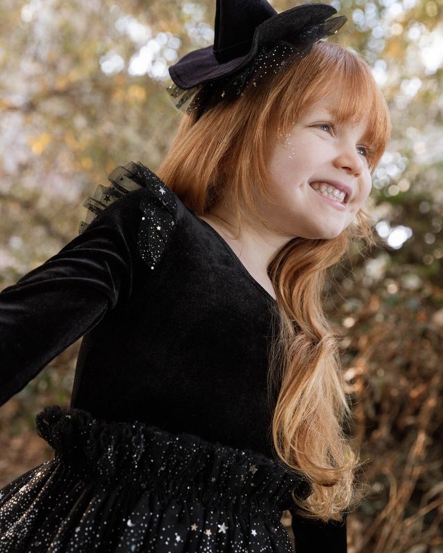 Child in a black witch costume with a hat, standing outdoors.