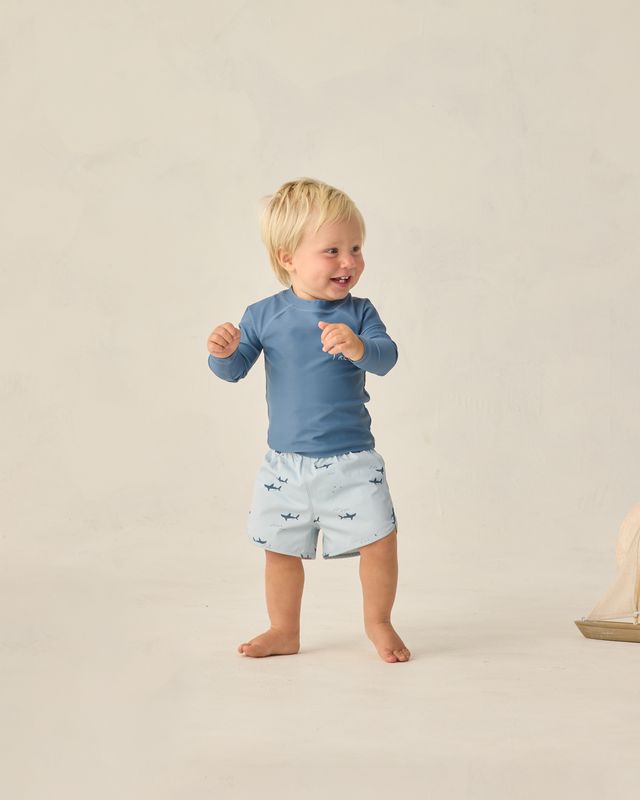 Child wearing a blue shirt and sharks patterned shorts on a beige background