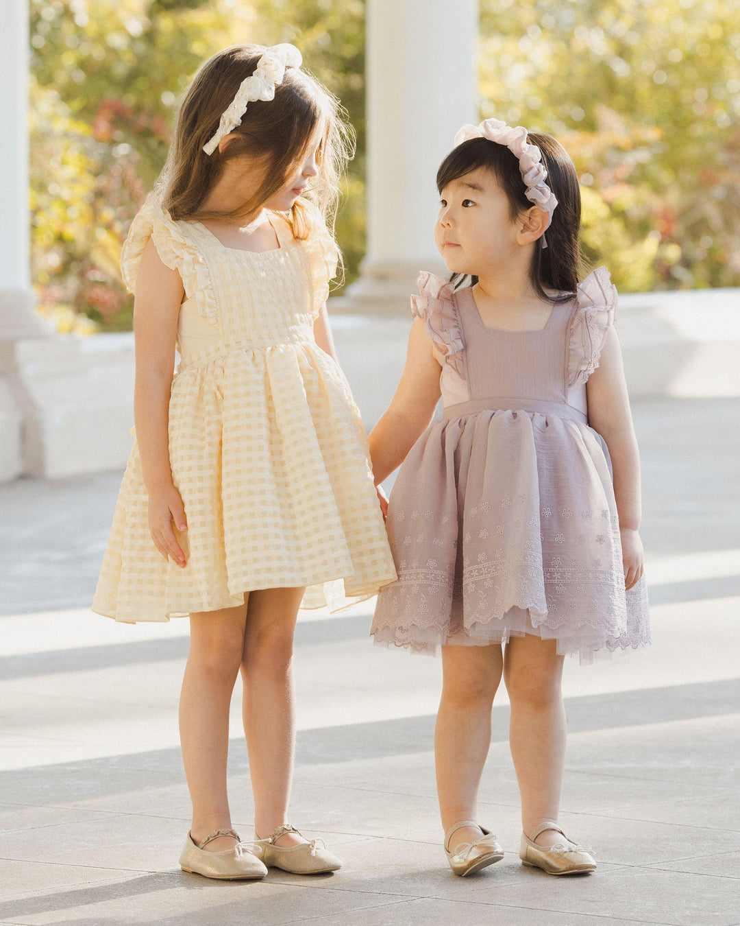 two little girls in the pinafore style holding hands, looking at one another. the dresses have a full skirt ruffle at the sleeve and bow in the back.