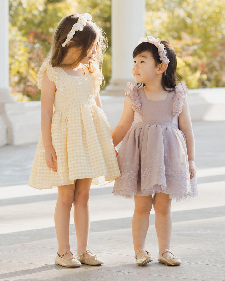 two little girls in the pinafore style holding hands, looking at one another. the dresses have a full skirt ruffle at the sleeve and bow in the back.