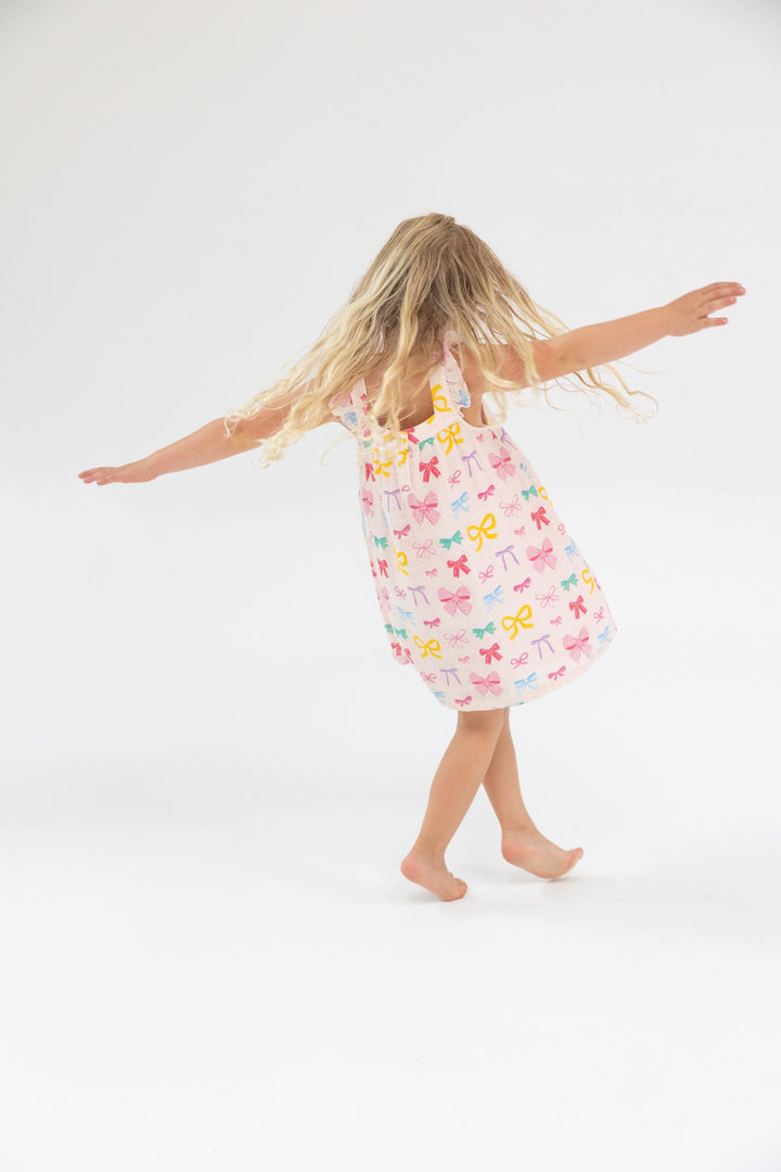 little girl modeling the a-line silhouette dress: white with multi colored bows: yellow, pink, green and blue. girl is twirling around so you can see the twirlability of this silhouette. 
