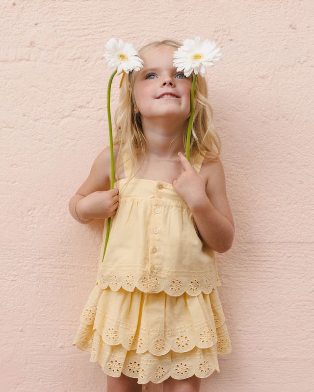 Young girl in a yellow two piece outfit with eyelet detailing holding flowers against a pink background