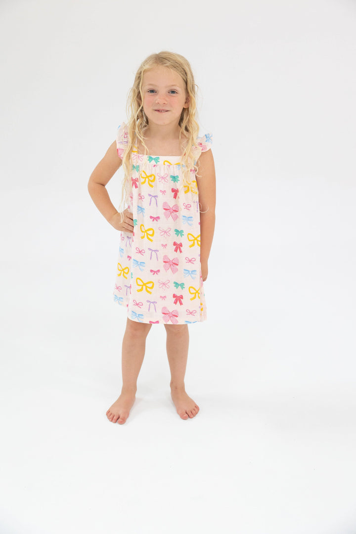 little girl wearing organic muslin cotton dress. a-line silhouette with ruffles at the sleeve. multi colored bows all over: pink, yellow, blue and green all over the dress
