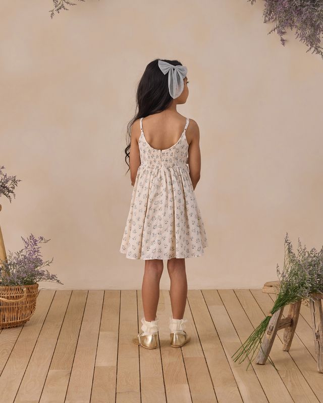 Young girl in a floral dress standing on a wooden floor with plants around.