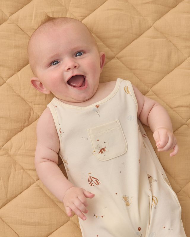 Baby wearing a cream-colored sleeveless romper with small cartoon carnival designs on a beige quilted surface