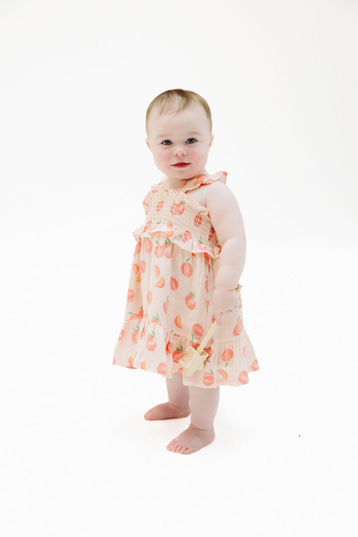 baby girl modeling the sweet peach dress. dress hits at the knee and has a smocked top. peaches and pieces of peaches all over the dress. 