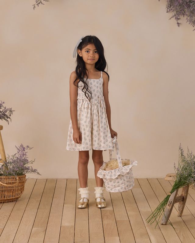 Young girl in a floral dress holding a basket, standing on a wooden floor with a beige wall and decorative plants in the background.