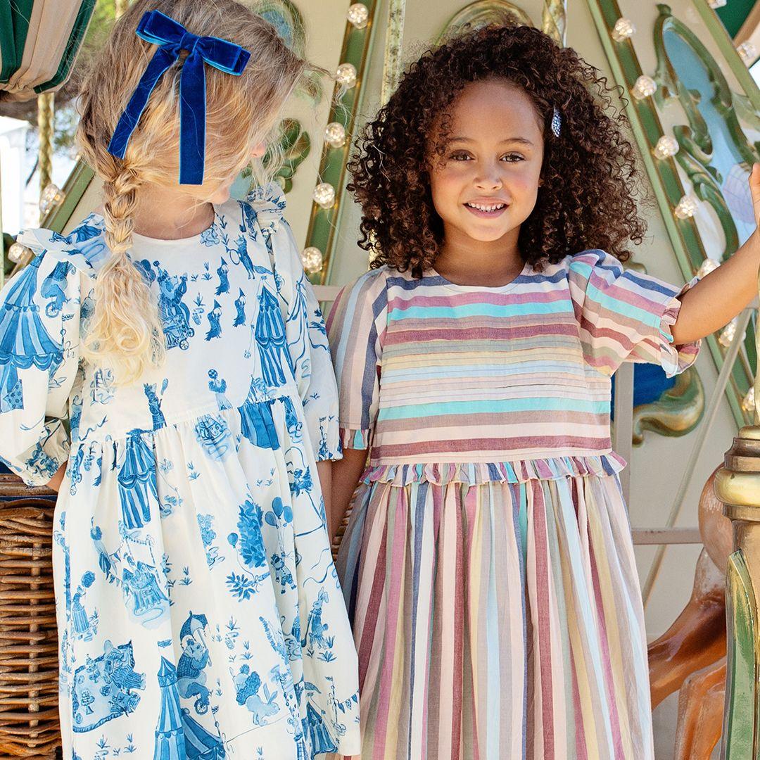 two young girls posing by the merry go around. one irl in a muted pastel striped dress. the other girl is wearing an ivory dress with blue circus print. 