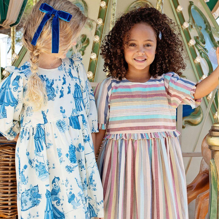 two young girls posing by the merry go around. one irl in a muted pastel striped dress. the other girl is wearing an ivory dress with blue circus print. 