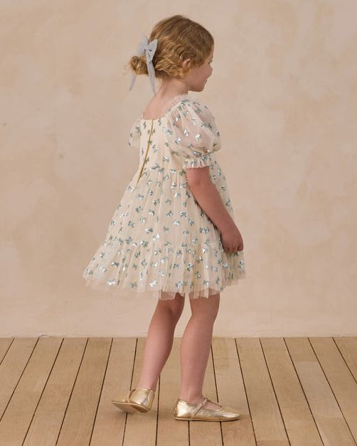 Young girl in a beige dress with blue sequins standing on a wooden floor with a beige wall background