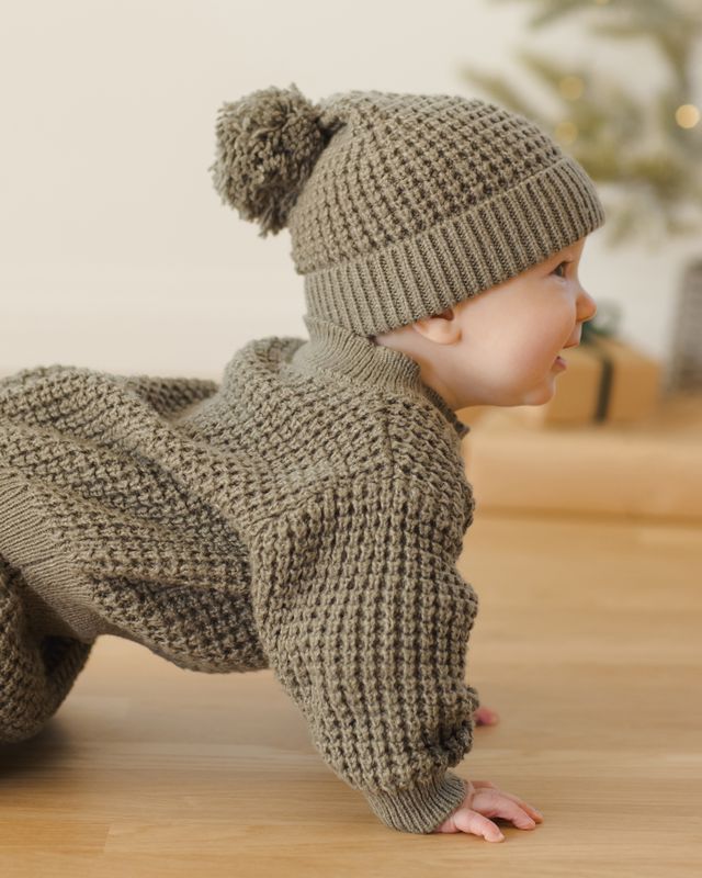Baby in a forest green knitted outfit and hat on a wooden floor.