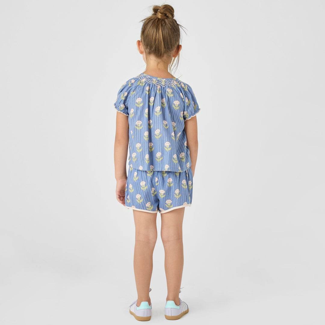 little girl modeling the periwinkle ava top and millie shorts set. this shows the top and shorts from the back. the top hits passed the waist and the shorts hit mid-thigh