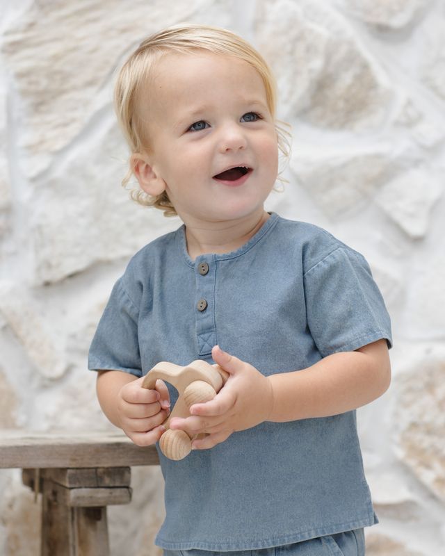 Child in a blue shirt holding a wooden toy against a textured white wall.