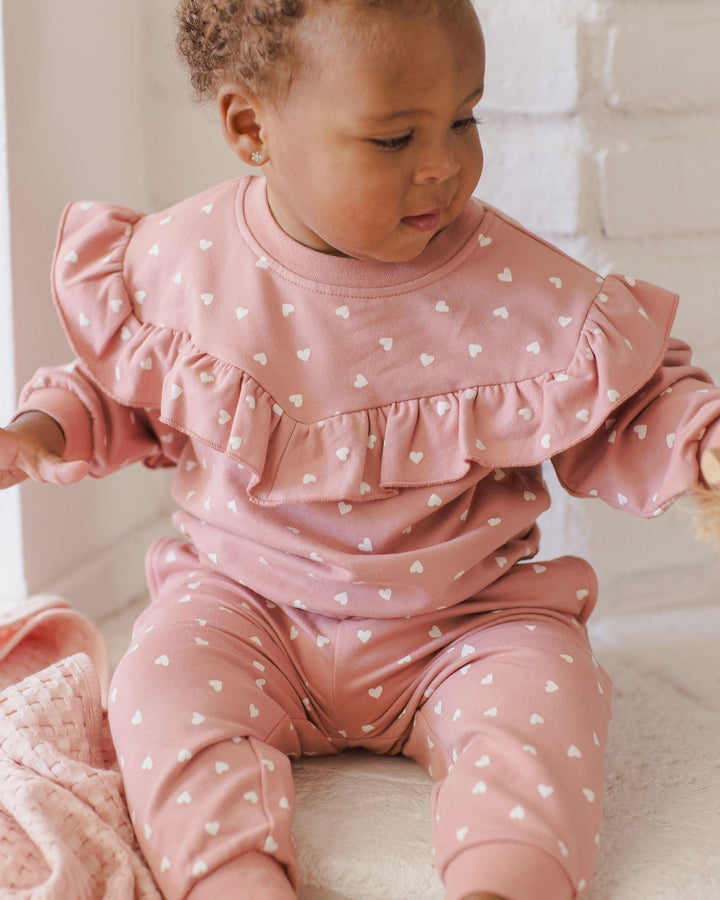 baby modeling the pink sweatsuit with tiny white hearts all over it. there is a ruffle that goes across the chest, from one arm to the other. 