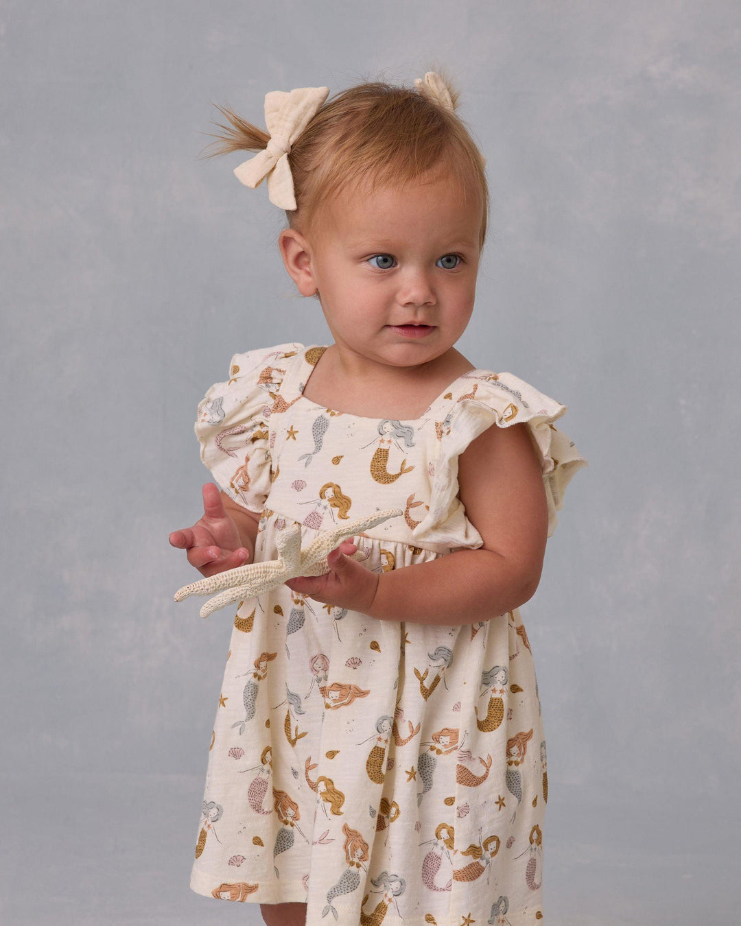 baby girl modeling the beige dress with mermaids all over. it shows you the length of the dress and how the sleeves are ruffled. 