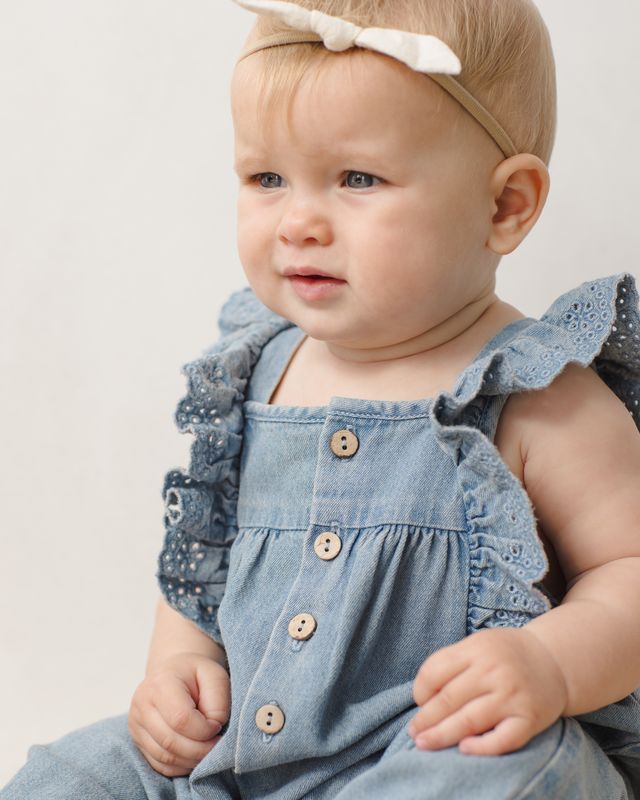 Baby wearing a blue denim jumpsuit with ruffles and buttons on a light background