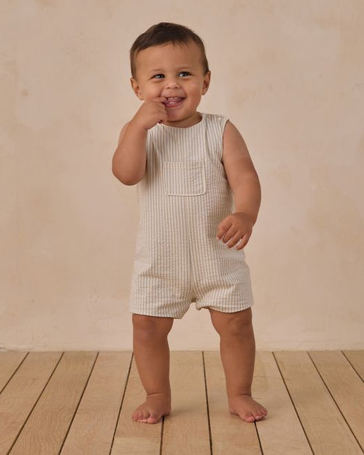 Baby in a striped romper standing on a wooden floor with a beige wall background