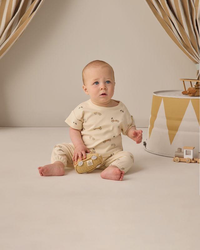 Baby sitting on a white floor wearing a beige outfit with small patterns, holding a toy.