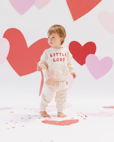 toddler boy modeling the sweatsuit with hearts in the background. The sweat set is beige, the top reads "Little Love" and the bottoms have a heart on one leg. 