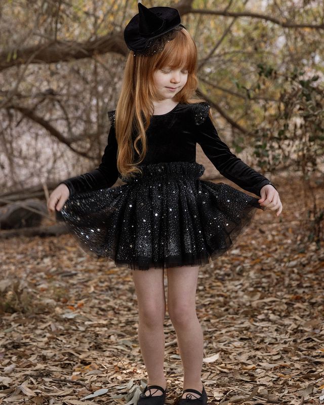 Young girl in a black dress with sparkles and a black hat standing in a forest.