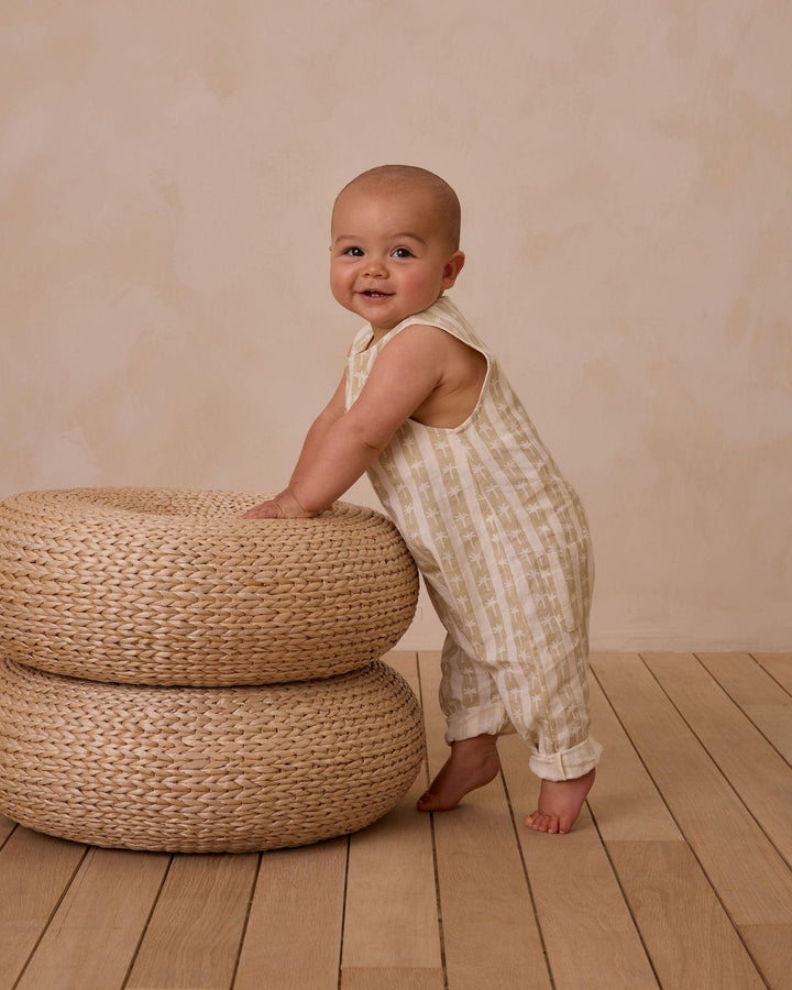 baby standing on the woven circle bench in the palm stripe jumpsuit. 