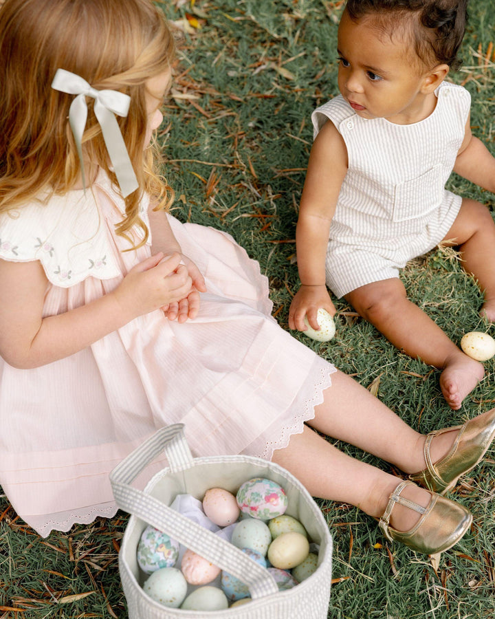 little girl sitting on the grass with a baby sharing eggs in a basket. the girl is dressed in a pink dress with a large collar. 