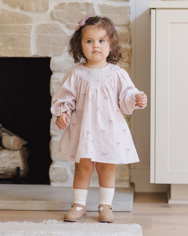 Young child in a light pink dress standing in front of a stone fireplace.