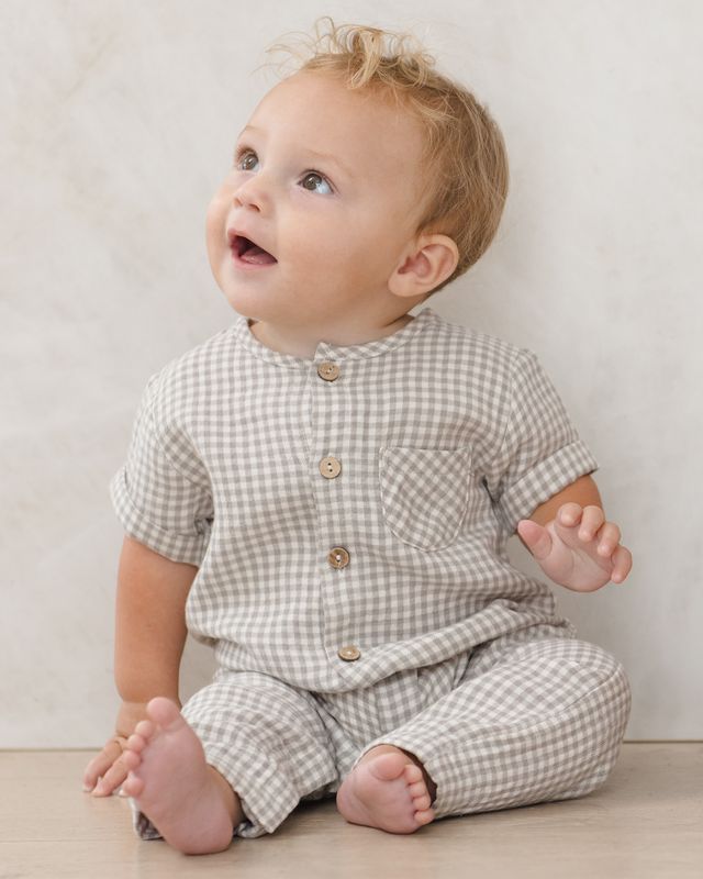 Baby wearing a sage checkered outfit sitting on a light wooden floor.
