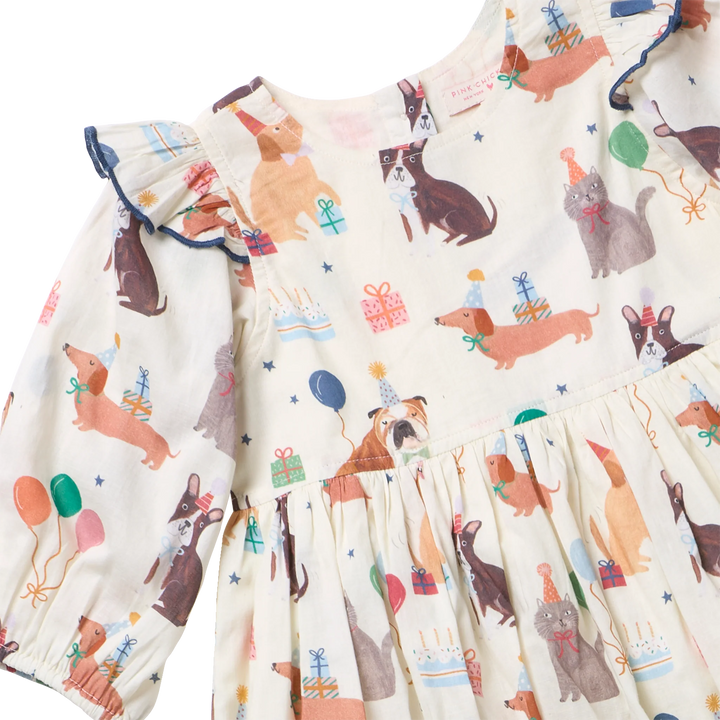 A close-up of a baby dress with a cream-colored base and an all-over print of dachshunds, birthday candles, party hats, balloons, and gift boxes. The dress features ruffled long sleeves with dark blue trim and a ruffled bodice and skirt.