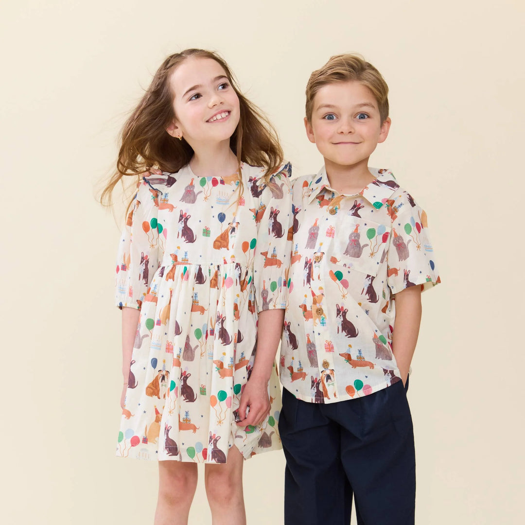 A young girl and boy modeling matching short-sleeve children's shirts and a dress with a colorful animal print on a cream background, set against a solid light beige backdrop.