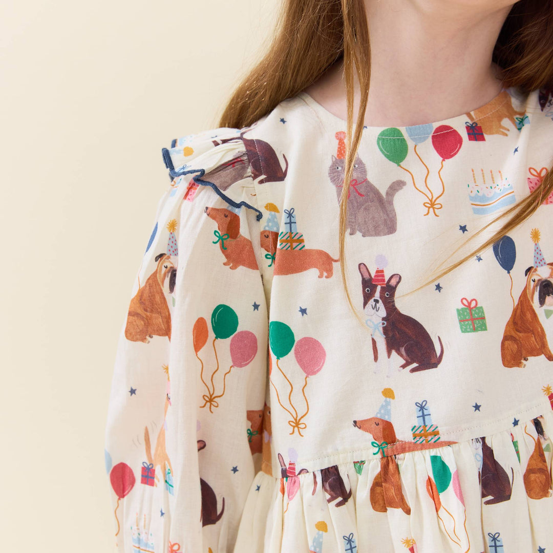 A close-up of a child wearing a cream-colored dress with a playful print of dogs, cats, balloons, gifts, and birthday candles. The dress features ruffled sleeves and a tiered skirt, shown against a plain light beige background.