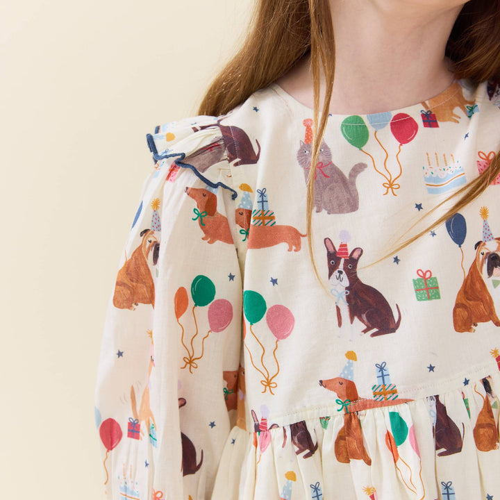 A close-up of a child wearing a cream-colored dress with a playful print of dogs, cats, balloons, gifts, and birthday candles. The dress features ruffled sleeves and a tiered skirt, shown against a plain light beige background.