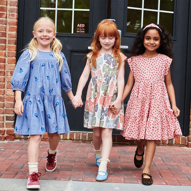 Three young girls holding hands and smiling, wearing colorful dresses in front of a building.