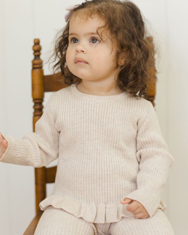 Child wearing a beige ribbed sweater sitting on a wooden chair.