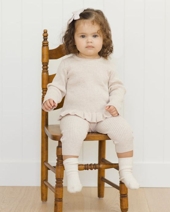 Child wearing a beige outfit sitting on a wooden chair against a white wall.