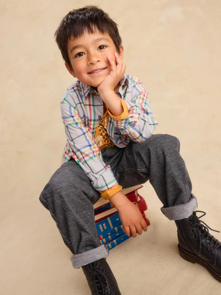 A young boy sitting on a stack of books, wearing a long-sleeve multicolor plaid shirt, dark gray cuffed pants, and black boots against a warm beige studio background.